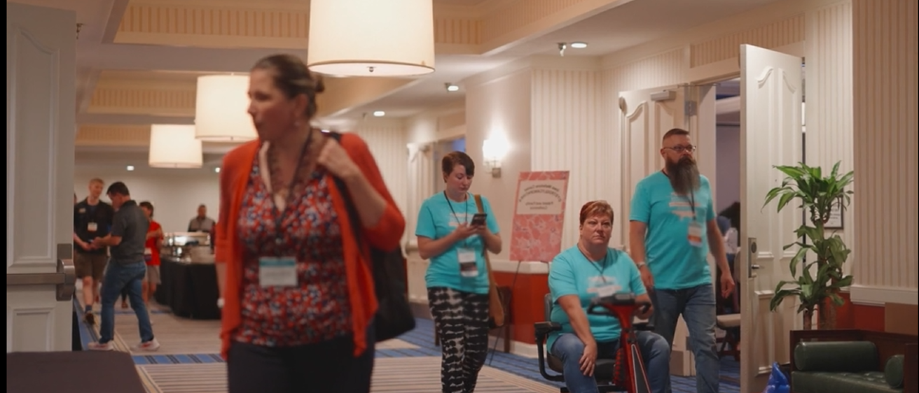 a screenshot from the linked video that shows 2 people walking and one riding an electric scooter wearing "2023 Wellstone Conference" shirts